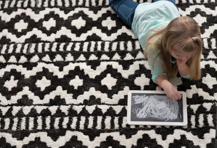 Young girl lying on a black and white patterned rug, drawing on a white-framed digital tablet with scribbles on the screen.