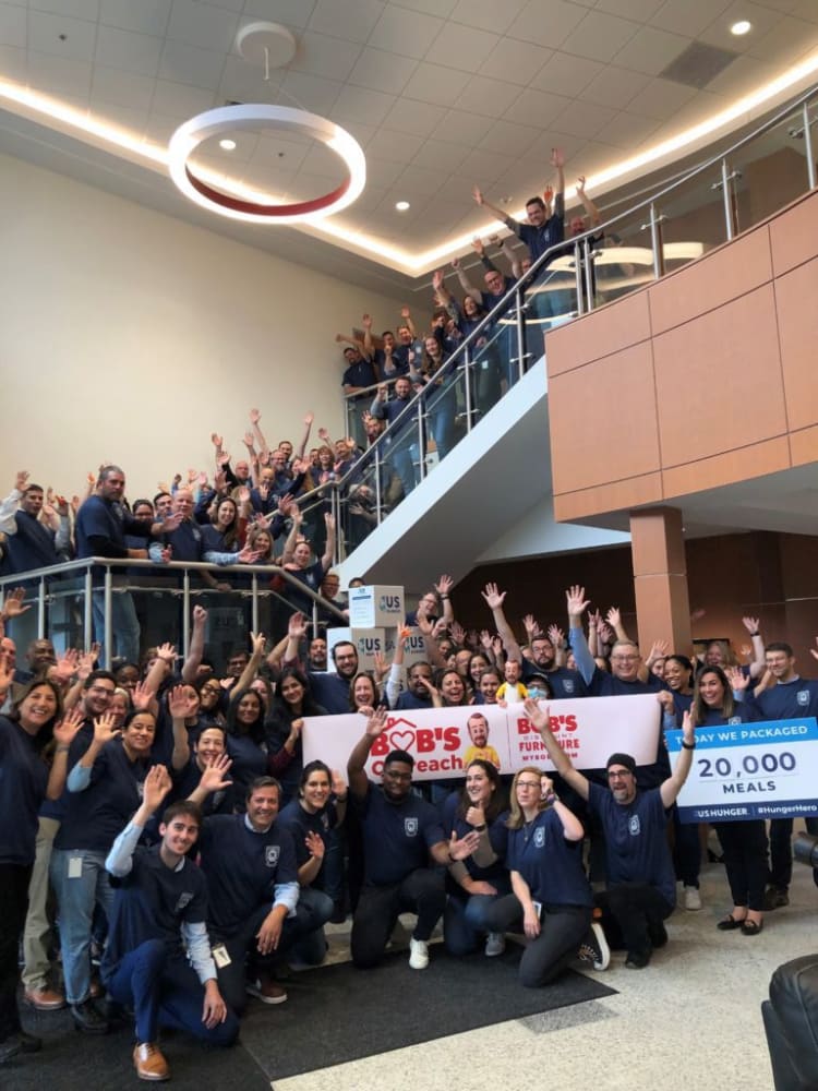 Large group of people in navy blue shirts gathered inside a modern building, holding banners that read "BOB'S Discount Furniture" and "Today We Packaged 20,000 Meals" for a community service event.