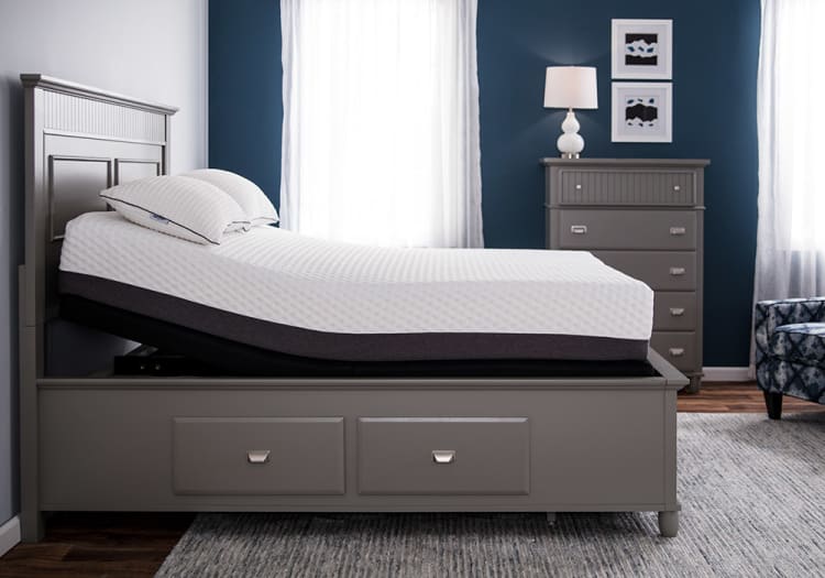 Adjustable gray wooden bed frame with white mattress and pillows, featuring storage drawers underneath, set in a modern bedroom with blue walls and matching dresser.