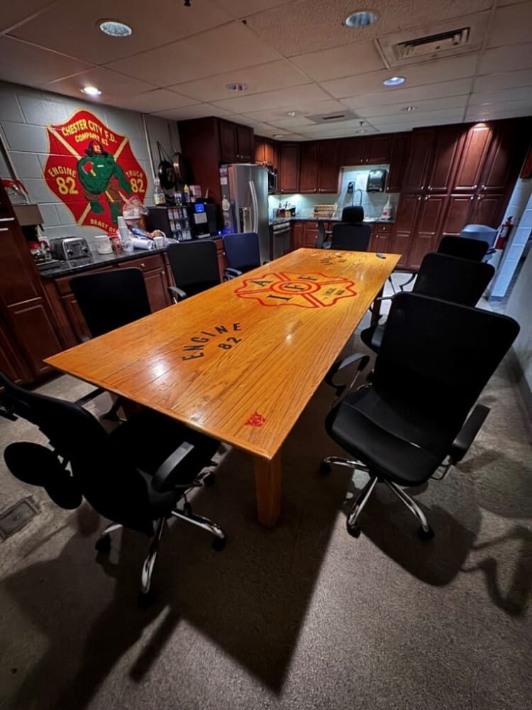 Conference room with a large wooden table featuring "ENGINE 82" and fire department logos, surrounded by black office chairs, in a kitchen setting.