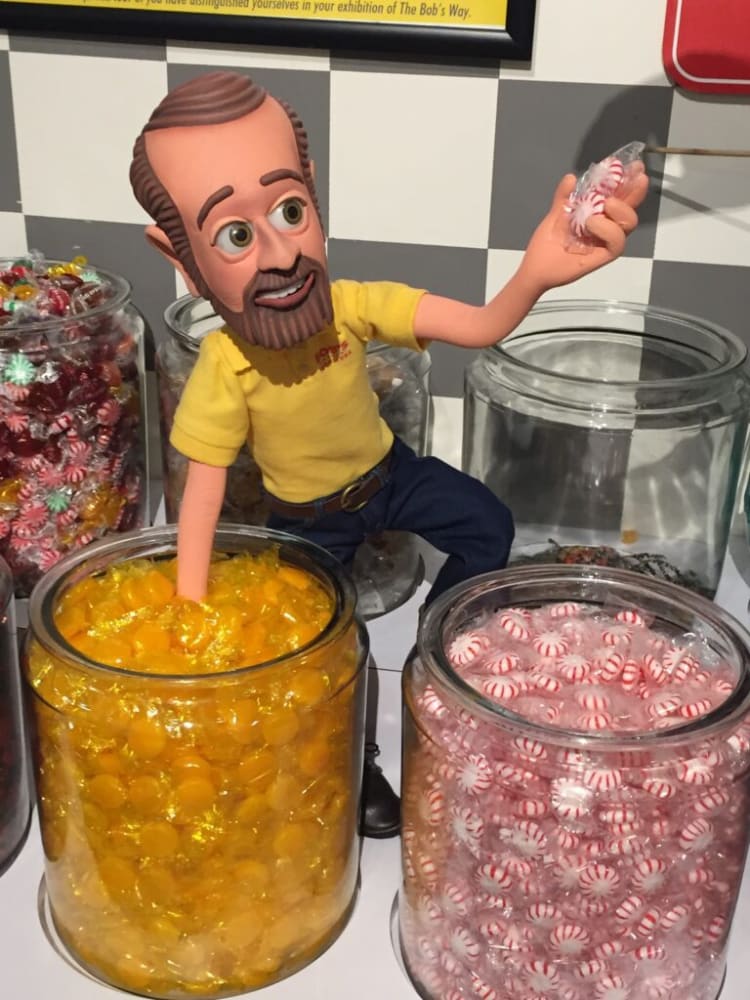 A candy display featuring a figurine with an arm dipped into a large glass jar filled with yellow hard candies, holding red and white striped peppermint candies above another jar filled with the same peppermint candies. The background has a black and white checkered pattern and additional jars of assorted colorful candies.