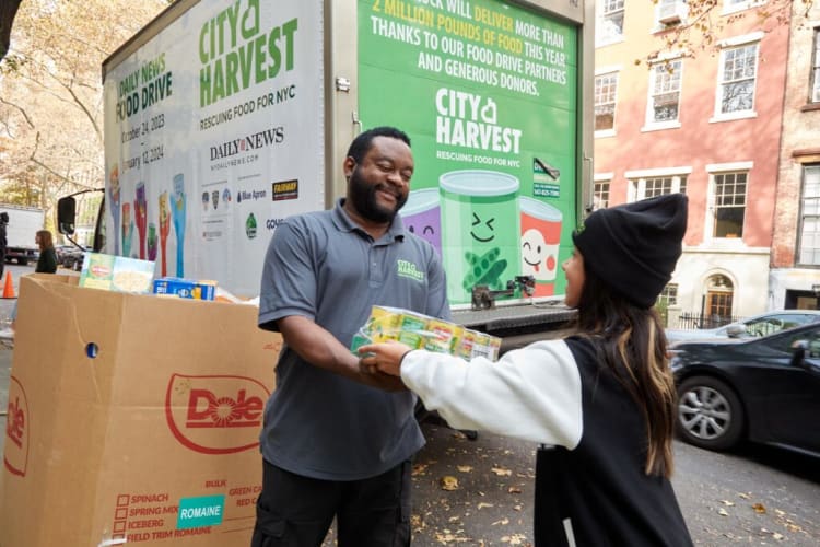 Two people exchanging packaged food items near a large City Harvest food drive truck with text about delivering 2 million pounds of food and donor thanks.