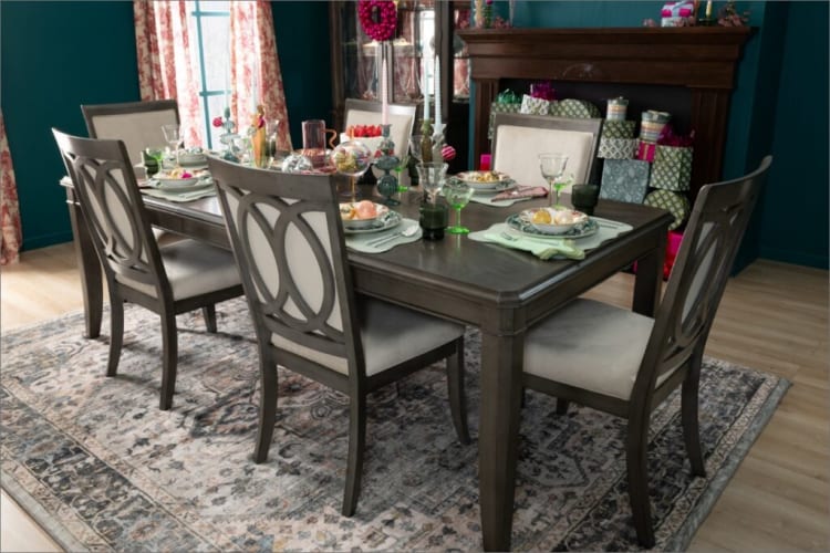 Dining room setup with a dark wood rectangular table and six matching chairs featuring cushioned beige seats and decorative backrests, set on a patterned area rug. The table is elegantly arranged with green glassware, patterned plates, pastel-colored napkins, and decorative ornaments, creating a festive and inviting atmosphere. Gift-wrapped presents are stacked near a dark wood fireplace mantel in the background, enhancing the holiday-themed decor.