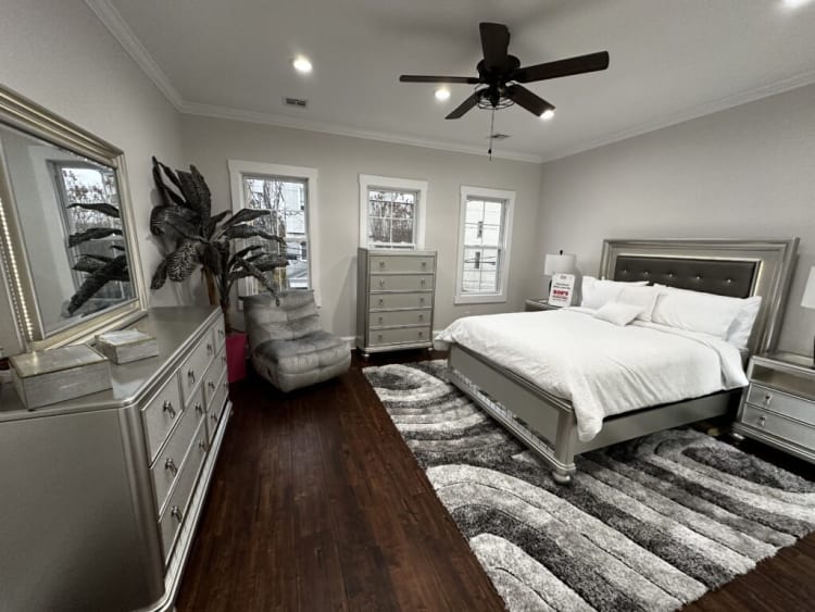 Modern bedroom with silver metallic furniture including a bed with white bedding, matching dressers, a gray armchair, dark wood flooring, and a black ceiling fan.