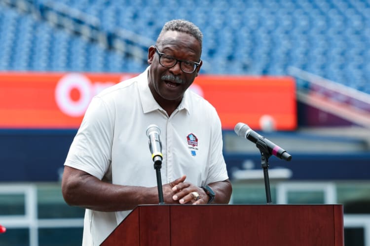 Man wearing a white short-sleeve polo with a Hall of Fame logo speaking at a wooden podium with dual microphones in a stadium setting.