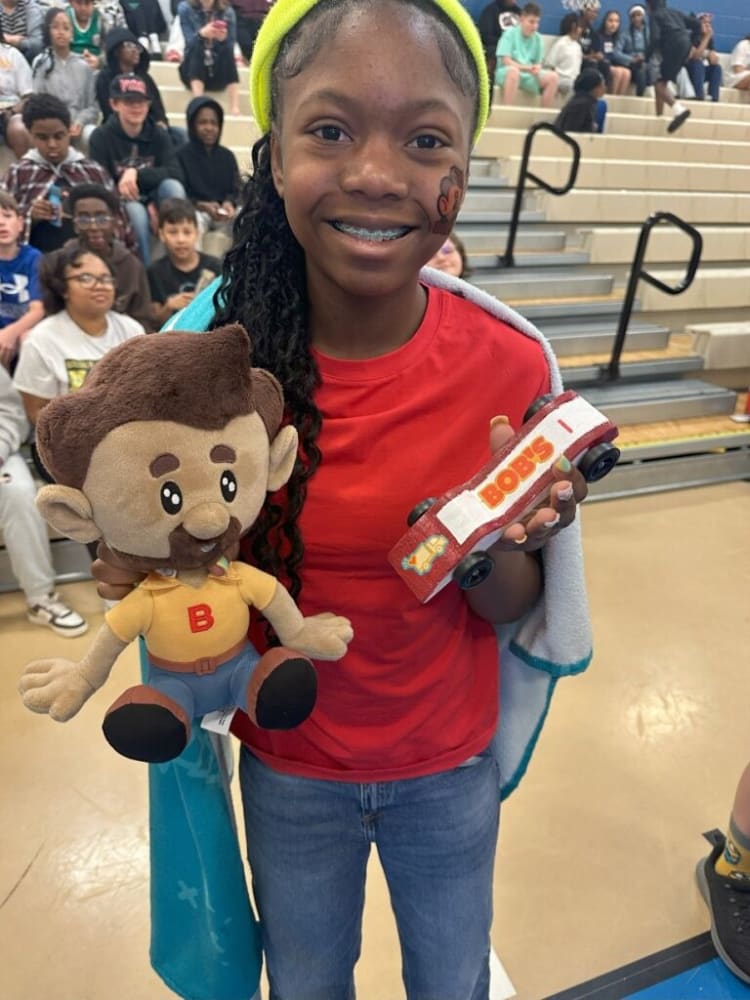 Person wearing a red shirt and jeans holding a plush toy with a "B" on its shirt and a red wooden car labeled "BOB'S 1" in a gymnasium setting.