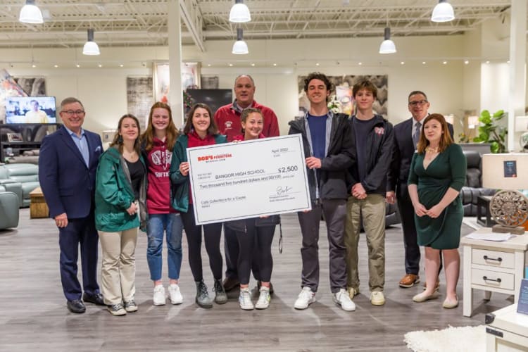 Group of people standing indoors holding a large check from Bob's Furniture for $2,500 made out to Bangor High School, dated April 2022.