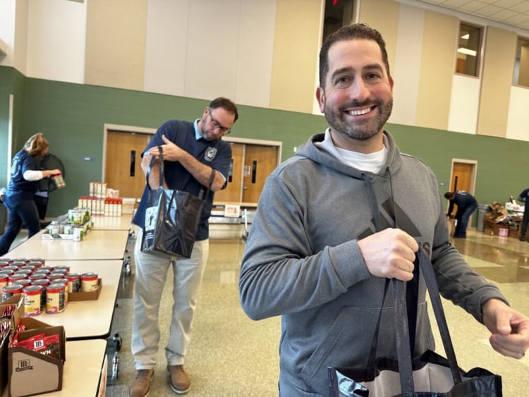 Two men holding black reusable shopping bags in a community food distribution setting with tables of canned goods and packaged food in the background.