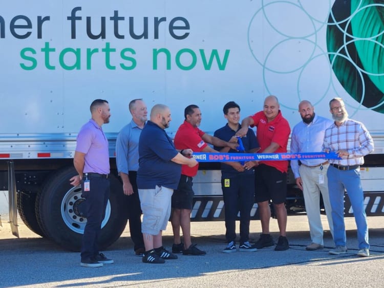 Group of men standing in front of a truck with the text "her future starts now," holding a blue ribbon that reads "Werner Bob's Furniture" during a ribbon-cutting event.