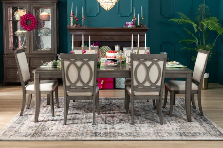 Dining room setup with a gray wooden table and six beige cushioned chairs on a patterned area rug, featuring a cake, candles, and decorative plants.