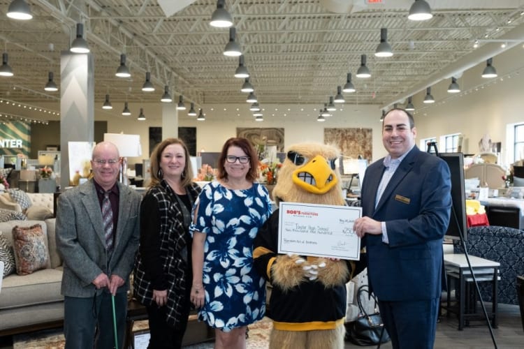 Five people, including a person in a yellow and black eagle mascot costume holding a large check from Bob's Furniture for $2,500 payable to Taylor High School, standing inside a furniture store. The check is dated May 21, 2023, and signed by Bob’s Furniture marketing manager, indicating a donation or sponsorship event. The store features sofas, lamps, and decorative pillows in the background.
