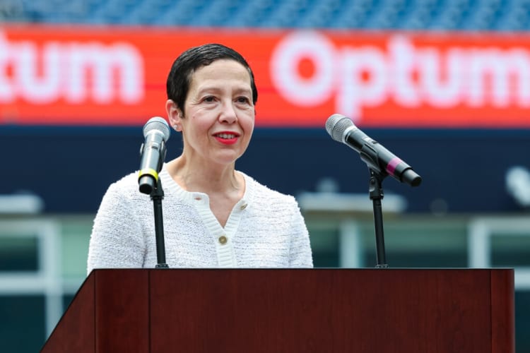 Person speaking at a podium with two microphones, wearing a white textured jacket, with a blurred red and white "Optum" sign in the background.