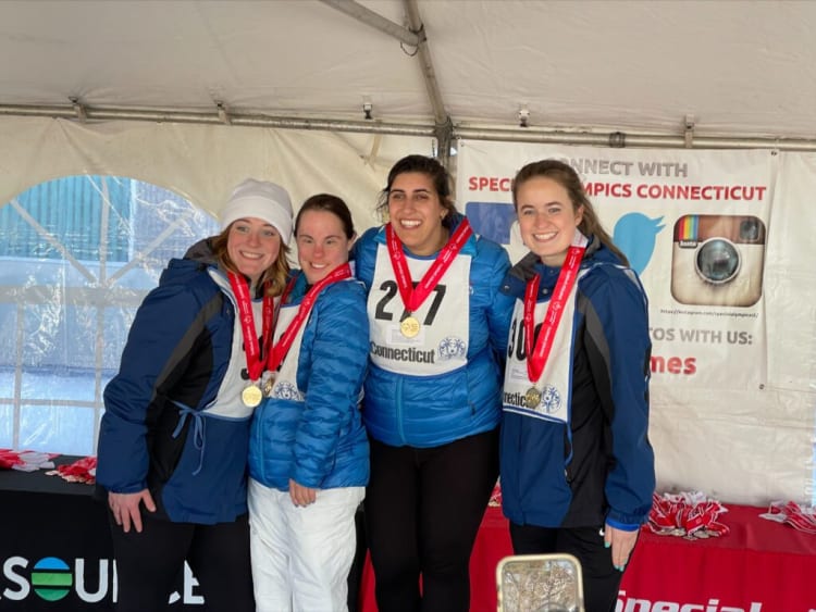 Four athletes wearing blue jackets and white bibs with "Connecticut" and Special Olympics logos, each with a red medal ribbon, stand under a tent. A banner behind them reads "CONNECT WITH SPECIAL OLYMPICS CONNECTICUT" with social media icons for Twitter and Instagram and the URL "https://instagram.com/specialolympicsct/". Medals are displayed on tables covered with black and red cloths.