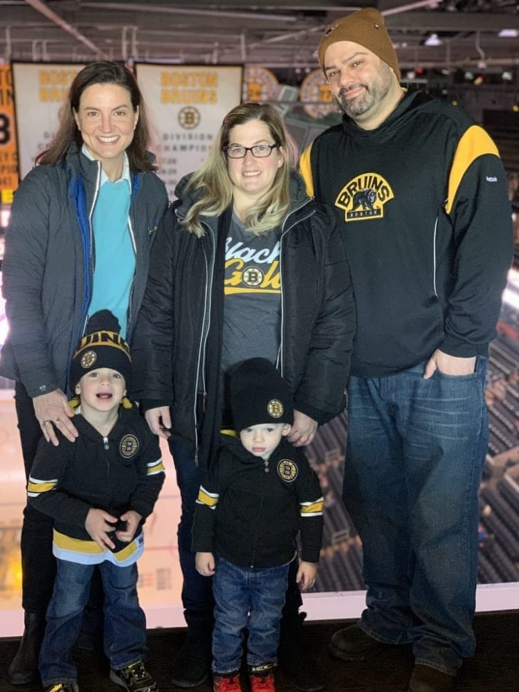 Family of five wearing Boston Bruins apparel, including black jackets, hoodies, and beanies with team logos, at an ice hockey arena.