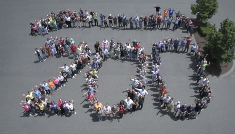 A large diverse group of people arranged outdoors to form the number "250," symbolizing a milestone or celebration, with trees and pavement visible.