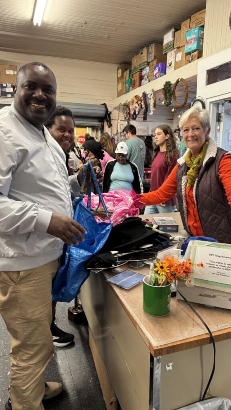 A person in a white jacket and beige pants holding a blue reusable shopping bag at a store counter with folded clothes and a green mug with orange flowers. A store employee wearing an orange shirt and brown vest is assisting. Shelves with boxes and items are visible in the background.