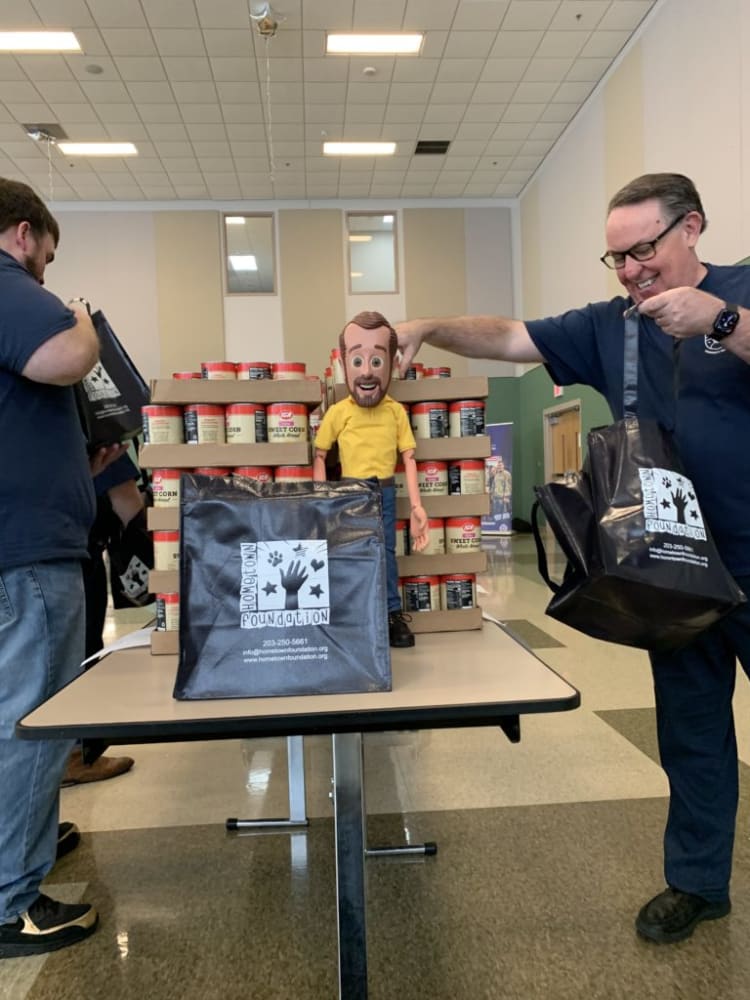 Volunteers distributing canned sweet corn from Hometown Foundation in black reusable bags with logo and contact info, inside a community center.