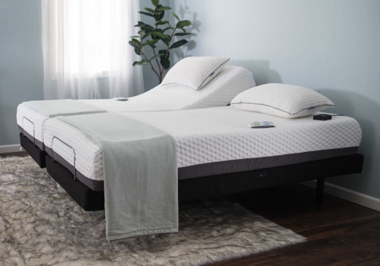 Adjustable split king bed with white quilted mattress covers, gray throw blankets, and matching pillows on a dark base, set on a plush rug.