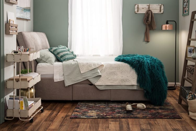 Modern bedroom with a tufted beige upholstered bed, teal textured throw blanket, patterned rug, wooden flooring, and soft green walls.