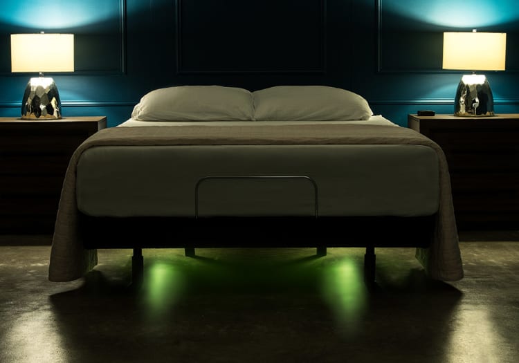 Modern bedroom with a king-sized bed featuring white bedding and a beige quilt, flanked by dark wood nightstands and metallic lamps, set against a teal paneled wall. Soft green under-bed lighting enhances the contemporary ambiance.