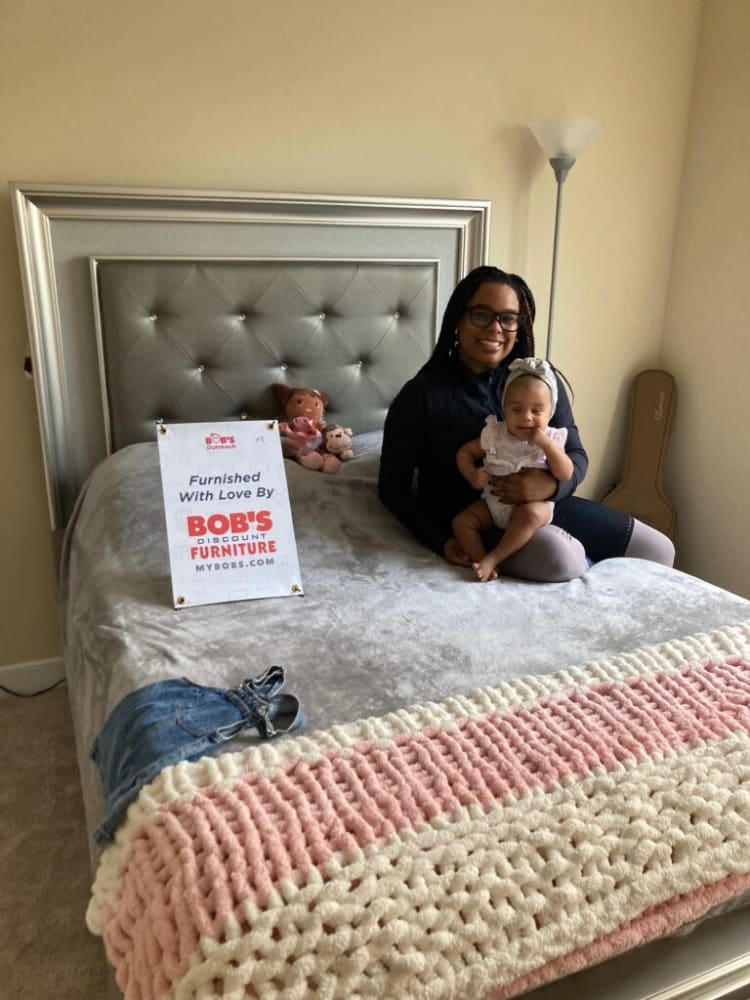 A modern silver-framed bed with a tufted gray headboard, soft gray bedding, a pink and cream chunky knit blanket, and a sign reading "Furnished With Love By BOB'S DISCOUNT FURNITURE MYBOBS.COM." A floor lamp and a guitar case are in the corner.