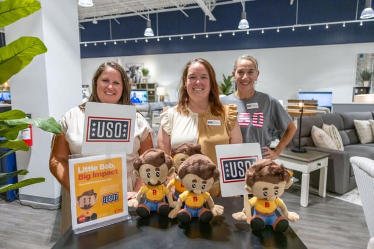 Three women stand behind a table with four plush dolls wearing yellow shirts and blue pants. Two signs display "USO" in bold letters. A flyer reads "Little Bob, Big Impact" promoting the USO organization. The setting appears to be an indoor event or store with modern furniture and plants in the background.