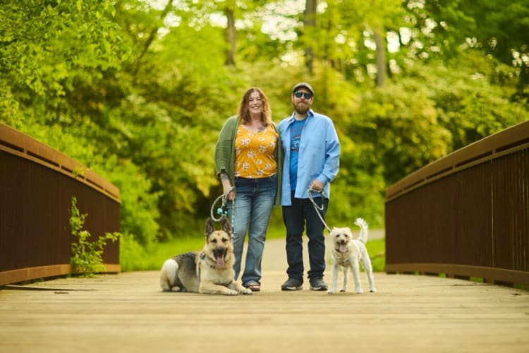 A couple standing on a wooden bridge surrounded by green trees, each holding a leash with their dogs—a large German Shepherd lying down and a small white dog standing.