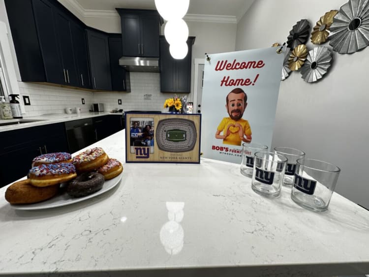 Modern kitchen countertop with a New York Giants framed stadium photo, "Welcome Home!" banner, and three clear glasses with NY Giants logo.
