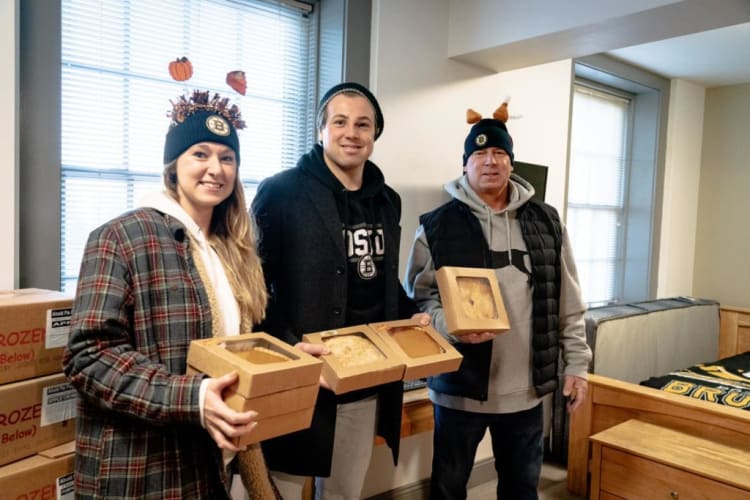 Three people wearing Boston Bruins winter hats and casual jackets hold multiple pumpkin pies in cardboard boxes inside a bright room with large windows.