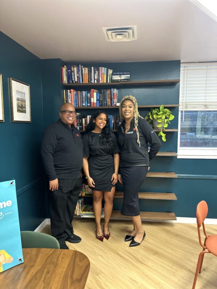 Three people standing in front of a dark teal wall with wooden shelves filled with books and a green potted plant, showcasing a professional office or library setting.