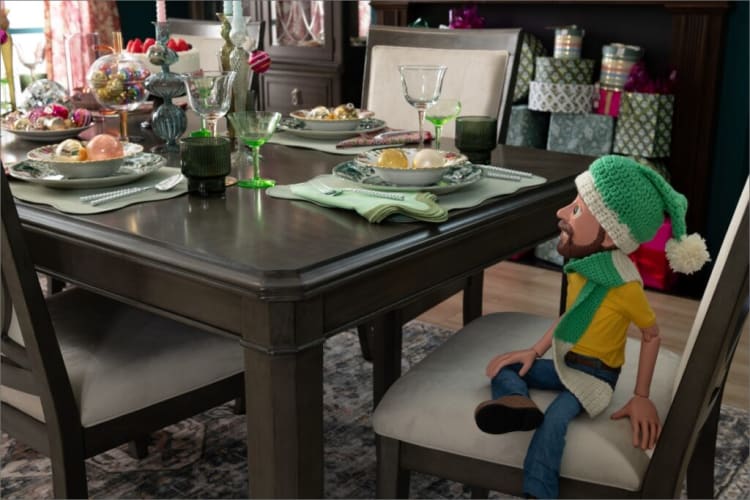 Wooden dining table set with green glassware, holiday-themed plates with ornaments, and a decorative doll wearing a green knit hat and scarf seated on a chair.