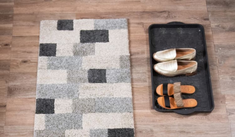 A geometric patterned gray and beige area rug next to a black plastic shoe tray holding beige slip-on shoes and brown slide sandals.