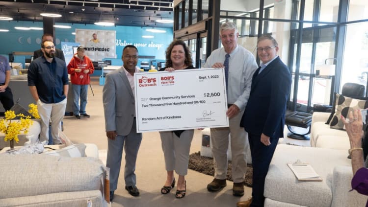 Five people stand in a furniture store holding a large check from Bob's Outreach for $2,500 to Orange Community Services, labeled "Random Act of Kindness." The store features modern sofas, cushions, and bright lighting.