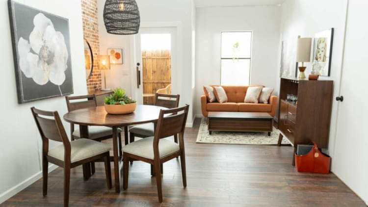 Modern dining and living area with wooden round table and four cushioned chairs, orange sofa, wooden coffee table, and decorative plants.