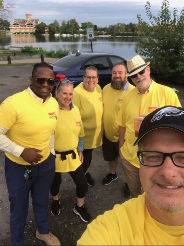 Group of six people wearing bright yellow Bob's Furniture branded T-shirts standing outdoors near a lake and parked car, some holding drinks.