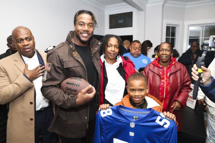 Group of people indoors, one holding a Wilson football, a child in front holding a blue New York Giants NFL jersey with number 58, casual and winter clothing.