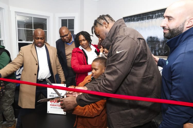 Group of people cutting a red ribbon at an indoor event, with a sign reading "Furnished With Love" and "Discount Furniture MYBODS.COM" on a black table.
