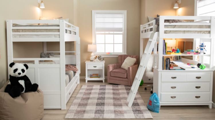 A modern kids' bedroom with white wooden bunk beds, one featuring a built-in desk and storage drawers, beige walls, a checkered area rug, a pink armchair, and soft lighting.