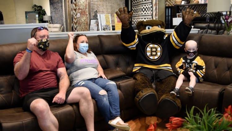 A family and a child wearing Boston Bruins hockey jerseys sitting on a brown leather sectional sofa with a mascot in a matching Bruins jersey raising its arms, in a living room setting with rugs and home decor in the background.