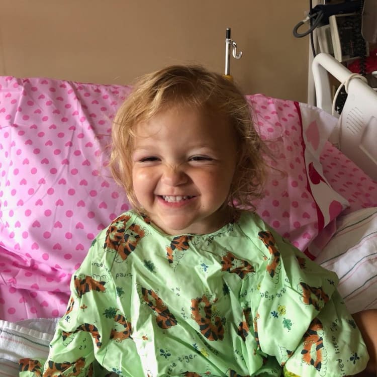 Child sitting on a hospital bed with pink heart-patterned sheets, wearing a light green hospital gown featuring tiger illustrations and text.