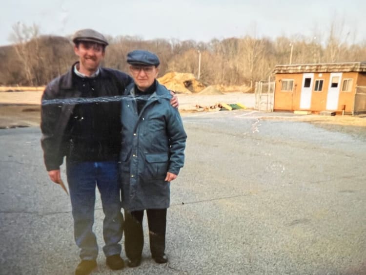 Two men standing on a paved outdoor area with bare trees and a small building in the background, wearing casual jackets and caps.