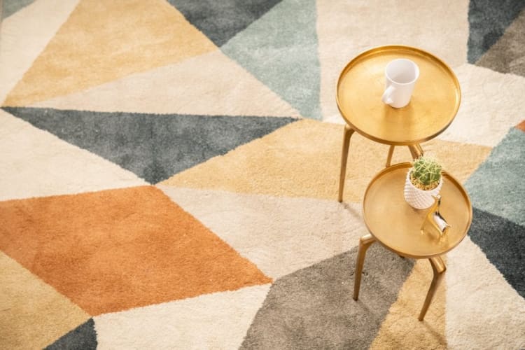 Two small round gold metal side tables on a geometric patterned rug with a white coffee mug on one and a small potted cactus with a gold bird figurine on the other.