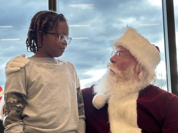 A child with braided hair wearing a gray sweatshirt with camouflage sleeves stands next to a person dressed as Santa Claus in a red suit with white fur trim, sitting by a large window with a cloudy sky outside.