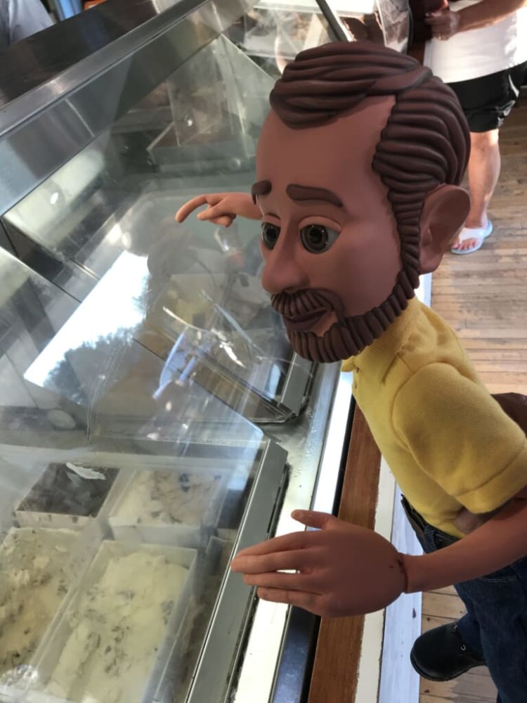 Child-sized mannequin with brown braided hair wearing a yellow shirt and jeans, pointing at ice cream flavors in a glass display case at an ice cream shop.