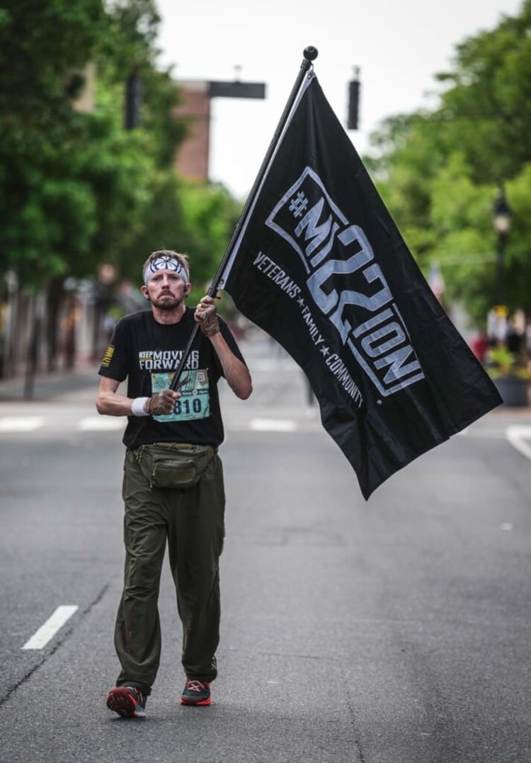 Man walking on street holding a black #M22tion flag with "Veterans * Family * Community" text, wearing black "Keep Moving Forward" shirt and olive pants.