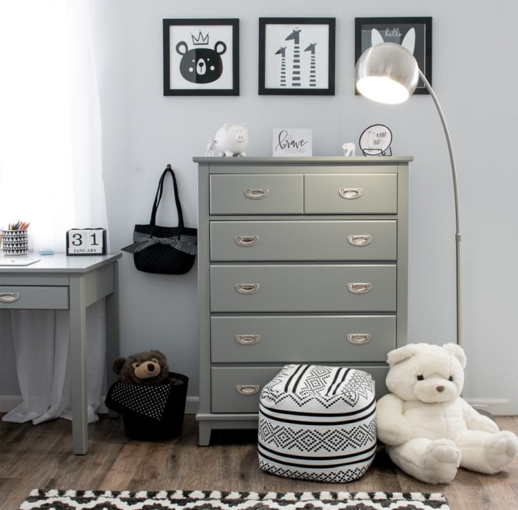 Gray wooden dresser with six drawers and silver handles in a modern nursery room, accompanied by a white plush teddy bear, patterned ottoman, and wall art.