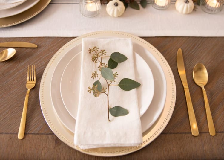 Elegant table setting with gold flatware, layered white plates, a beige beaded charger, and a white linen napkin adorned with a green eucalyptus sprig.