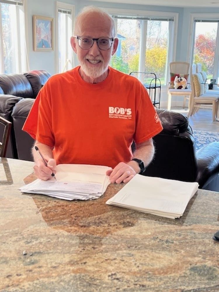 Person wearing a red Bob's Discount Furniture, Inc. t-shirt writing on a stack of papers at a granite countertop in a bright living room.