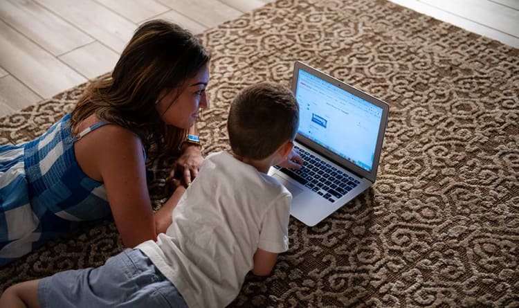 Woman and child lying on a patterned brown carpet using a silver laptop with a webpage open, engaging in online learning or browsing.