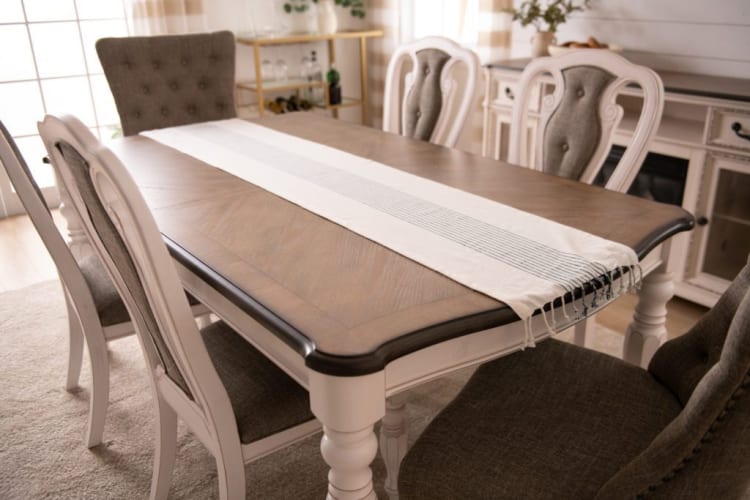 Dining table set with a wooden top and white painted legs, featuring six upholstered chairs in gray fabric and a white table runner with black stripes.